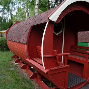 Rotes Holzfass mit überdachtem Sitzbereich im Grünen, ideal für Campingurlaub.