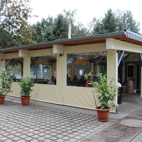 Überdachte Terrasse eines Restaurants mit großen Fenstern und Pflanzen in Töpfen vor dem Eingang.