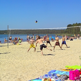 Junge Menschen spielen Beachvolleyball am sonnigen Sandstrand mit See und Badegästen.
