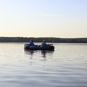 Zwei Personen rudern in einem Schlauchboot auf einem ruhigen See bei sonnigem Wetter.